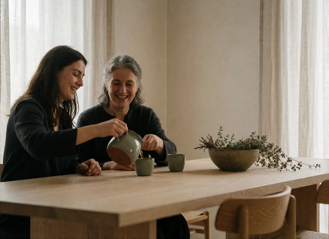 Two Armenian women laughing together while pouring tea at a long pale oak dining table.